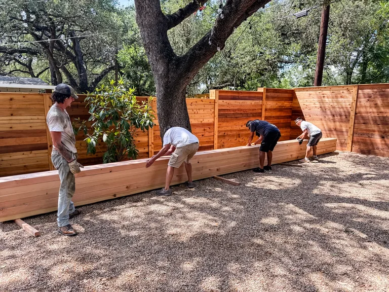 Four crew members carrying a long timber board through a fenced courtyard beneath a tree